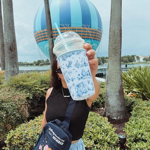 Könnte beinhalten: Ein klarer Plastikbecher mit blauem Strohhalm und einer weiß-blauen Hülle mit Cartoonfiguren. Eine marineblaue Umhängetasche mit dem Text "Disney Vacation Club Member" und einer Cartoon-Grafik ist ebenfalls sichtbar.