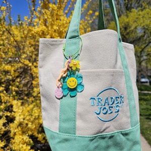 May include: A canvas tote bag with a light gray body and mint green accents, including the handles and a pocket. The bag features the Trader Joe's logo in blue embroidery and is adorned with a charm featuring smiley face and flower designs.