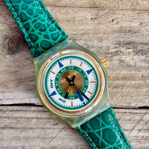 Op de afbeelding: Een vintage Swatch-horloge met een groen en gouden wijzerplaat en een bijpassende groene band met textuur. De wijzerplaat heeft witte, blauwe en gouden accenten met de woorden "OFF" en "ON". Het horloge staat tegen een houten achtergrond.