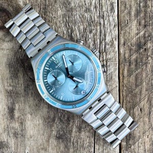 May include: A Swatch chronograph watch with a light blue dial and bezel, featuring black hands and silver-tone hour markers. The watch has a stainless steel band and is set against a wooden background. The watch face displays the Swatch logo.