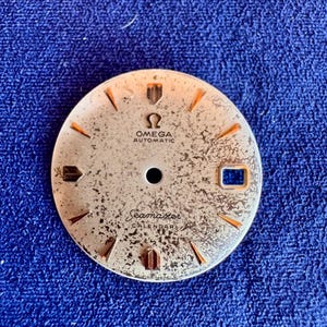 Peut inclure: Un cadran de montre rond de couleur argentée avec une surface texturée et patinée. Il affiche le logo Omega, le texte OMEGA AUTOMATIC et Seamaster CALENDAR. Le cadran comprend des index dorés, un guichet de date rectangulaire à trois heures et la mention SWISS MADE en bas.