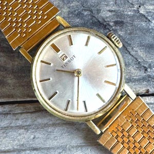 May include: A gold-toned Tissot wristwatch with a silver face and gold hands. The watch features a gold band with a woven design. The watch face has gold markers and the Tissot logo.