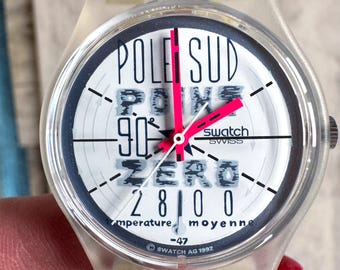 Può includere: Un orologio Swatch trasparente con quadrante bianco con le parole "POLE SUD", "ZERO" e "90°". L'orologio ha una lunetta nera, lancette rosa e la scritta "SWISS". Il cinturino dell'orologio è trasparente.