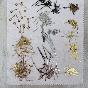 May include: A collection of various watch hands in gold, silver, and black. The hands are in different styles and sizes, with pointed and circular designs. The hands are scattered on a light gray surface.