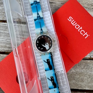 May include: A Swatch watch with a black face and a strap featuring images of the Eiffel Tower and silhouettes. The watch is in a clear plastic case next to a red Swatch box.