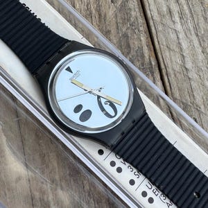 May include: A Swatch wristwatch featuring a black ribbed silicone strap and a round black case. The watch face is silver with black circular and oval graphic accents, silver hands with gold-tone accents, and the Swatch Swiss logo at the top.