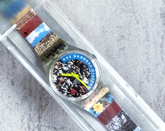 Può includere: Un orologio in plastica trasparente con un cinturino fantasia con varie immagini e colori. Il quadrante dell'orologio ha la scritta "ONE HUNDRED MILLION" in blu e una scena di folla. L'orologio è in una custodia di plastica trasparente.
