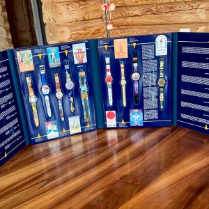 Puede incluir: Una colección de ocho relojes de pulsera Swatch presentados en un estuche azul. Cada reloj presenta diseños gráficos y colores únicos, acompañados de pequeñas tarjetas impresas y textos que detallan la historia de varios Juegos Olímpicos.