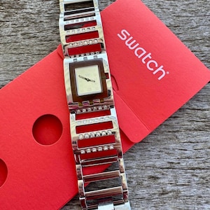 May include: A silver watch with a square face and a brown dial. The watch has a silver band with rhinestones. The watch is on a red background with the word "swatch" printed on it.