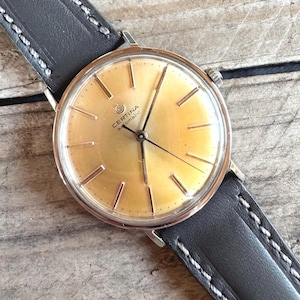 Può includere: Un orologio automatico Certina vintage con quadrante dorato e cassa argentata. L'orologio ha un cinturino in pelle nera con cuciture bianche. Il quadrante presenta indici a bastoncino e il logo Certina.