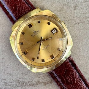May include: A gold-toned Fortis Tuxedo automatic wristwatch with a rectangular face and a brown leather band. The watch face displays the date and the word "Tuxedo". The watch has a gold-colored bezel and a brown leather strap.