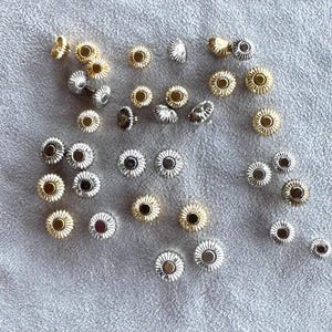 May include: A collection of watch crowns in gold and silver tones. The crowns have a fluted, flower-like design with a circular center. The crowns are scattered on a light gray surface.