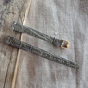 May include: Two gray watch straps with a crocodile pattern. One strap has a gold-colored buckle. The straps are made of a textured material and are displayed on a neutral background.