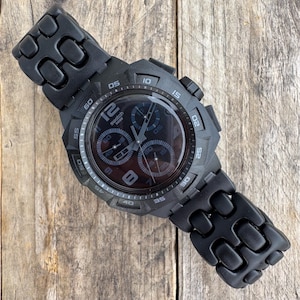 Könnte beinhalten: Eine mattschwarze Swatch-Chronographenuhr mit rundem Gehäuse, strukturiertem Gliederarmband und einem dunklen Zifferblatt mit drei Hilfszifferblättern. Die Lünette zeigt Minutenmarkierungen in Fünferschritten und das Zifferblatt trägt das Swatch-Logo.