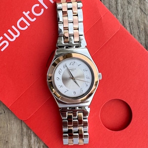May include: A Swatch wristwatch featuring a white dial with a circular textured center, black Arabic numerals, and a date window at the six o'clock position. The watch has a two-tone metal link bracelet in silver and rose gold, with a matching bezel.