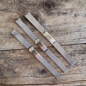 May include: Three silver-toned metal watch bands featuring a fine mesh texture. Each band includes a smooth, rectangular metal clasp engraved with a small horseshoe-shaped logo. The bands are arranged diagonally, showing a consistent design.