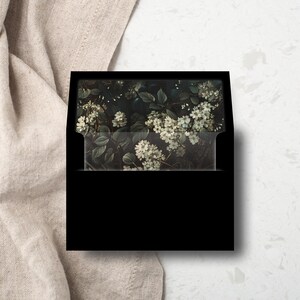 Peut inclure: Une enveloppe noire avec un motif floral composé de fleurs blanches et de feuilles vertes. L'enveloppe est fermée et possède un rabat.