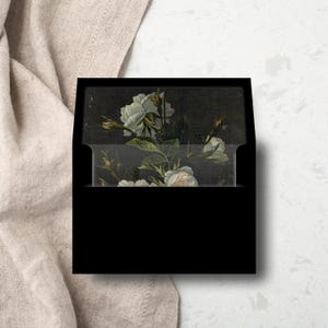 May include: Black envelope with a white floral design on the front. The flowers are white roses with green leaves and stems. The background is a dark, almost black, color.