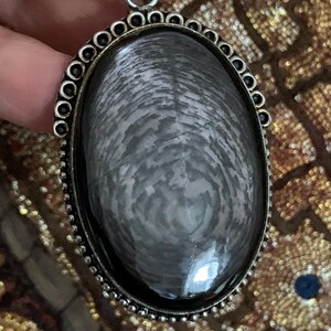 May include: A silver oval pendant with a black and grey stone featuring a swirling pattern. The pendant is surrounded by a decorative silver border.