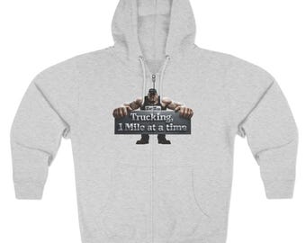 May include: A light grey zip-up hoodie featuring a graphic of a muscular figure wearing a cap and dark clothing, holding a rectangular sign that reads "Trucking, 1 Mile at a time" in white text. The hoodie includes a hood with drawstrings.