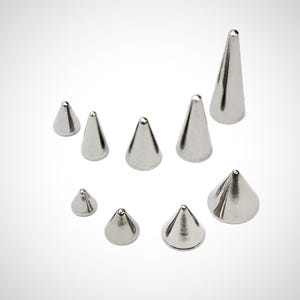 Puede incluir: Un conjunto de nueve tachuelas de cono de plata de diferentes tamaños. Las tachuelas están hechas de metal y tienen un acabado liso y pulido.