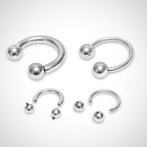 Könnte beinhalten: Zwei silberne hufeisenförmige Ringe mit Kugelenden und zwei zusätzliche Kugelenden. Die Ringe sind unterschiedlich groß.