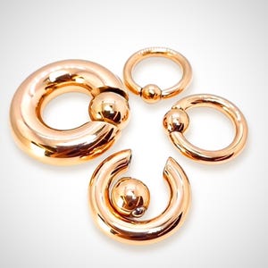 Puede incluir: Cuatro anillos de joyería corporal de metal de color dorado con cierres de bola. Los anillos son de diferentes tamaños y están dispuestos en un semicírculo.