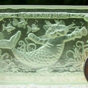 May include: A clear glass paperweight with a raised design of a cat with a mermaid tail. The cat is swimming in the ocean with fish and seaweed. The text "Sam the Kitty Hawk" is inscribed on the top of the paperweight.