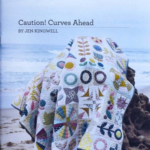 Könnte beinhalten: Ein farbenfroher Quilt mit einer Vielzahl von geometrischen Formen und Mustern, darunter Kreise, Quadrate und Dreiecke. Der Quilt ist gefaltet und über einen Felsen an einem Strand gelegt. Der Text "Caution! Curves Ahead" und "By Jen Kingwell" ist auf dem Cover gedruckt.