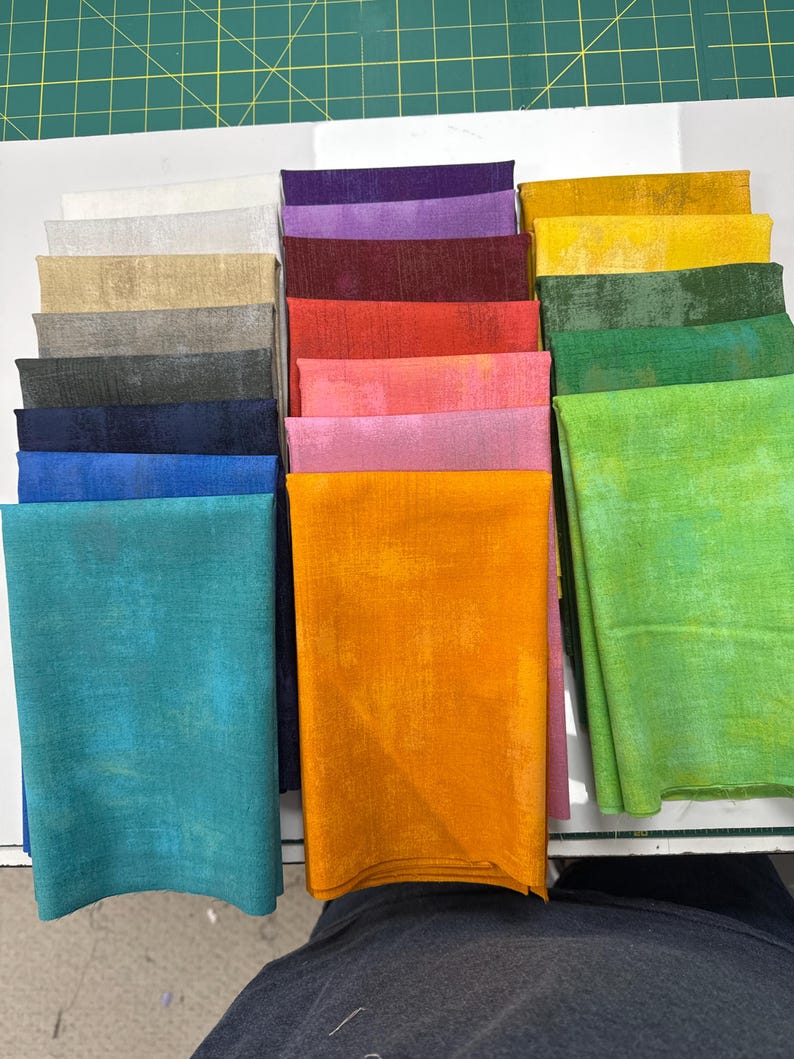 May include: A collection of folded fabric squares arranged in three vertical columns. The fabric features a mottled, textured appearance in a wide spectrum of colors including white, gray, black, blue, teal, purple, red, pink, orange, yellow, and green.