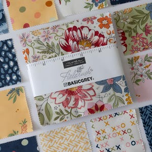 Peut inclure: Un assortiment de carrés de tissu avec divers motifs floraux et géométriques. Le paquet central, enveloppé d'une bande blanche, porte l'inscription "Fieldrush" et "BASICGREY". Les carrés présentent une gamme de couleurs, dont le rose, le bleu, le jaune et le vert.