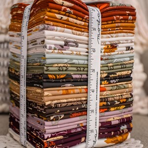 May include: A stack of fabric bundles in various colors, including orange, brown, and green, tied with a white ribbon printed with "moda fabrics." The fabric appears to be patterned with floral designs. The ribbon shows measurements in inches.