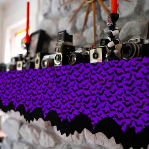 May include: A Halloween-themed display featuring a purple and black bat-patterned table runner. Vintage cameras and decorative candles sit atop a stone mantel. A starburst clock is visible in the background.