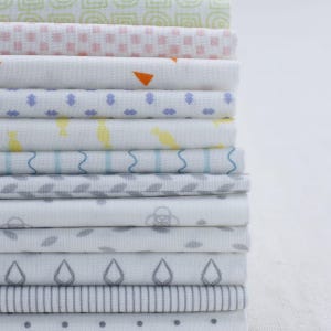 May include: A stack of 12 fabric squares with different patterns in shades of white, gray, blue, and pink. The patterns include geometric shapes, stripes, and dots.
