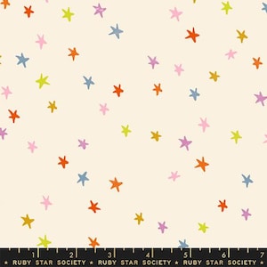 以下が含まれることがあります： オレンジ、ピンク、イエロー、ブルーの小さな手描き風の星が散りばめられたクリーム色の生地。下部にある黒い定規はセンチメートル単位の目盛りを表示しており、生地の幅が18cmであることを示しています。