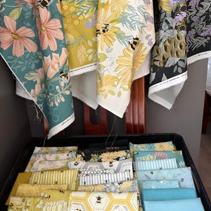 Puede incluir: Surtido de piezas de tela con estampados florales y de abejas en una variedad de colores, incluyendo turquesa, amarillo y negro. Las telas se exhiben en una bandeja negra y colgadas de una silla de madera.