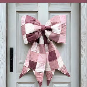 Op de afbeelding: Een grote, gewatteerde strik in roze en bordeaux tinten hangt aan een witte deur. De strik is gemaakt van vierkante stof en heeft lange, puntige uiteinden. Tekst op de afbeelding luidt "THE BIG GIANT QUILTED BOW" en "Designed to be reversible".