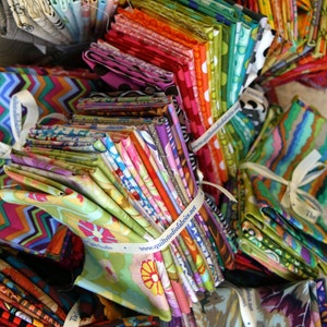 May include: A large stack of colorful fabric scraps with various patterns and designs. The fabrics are arranged in a pile and tied with white ribbon. The ribbon has the text "www.quiltsudiofabrics.net" printed on it.