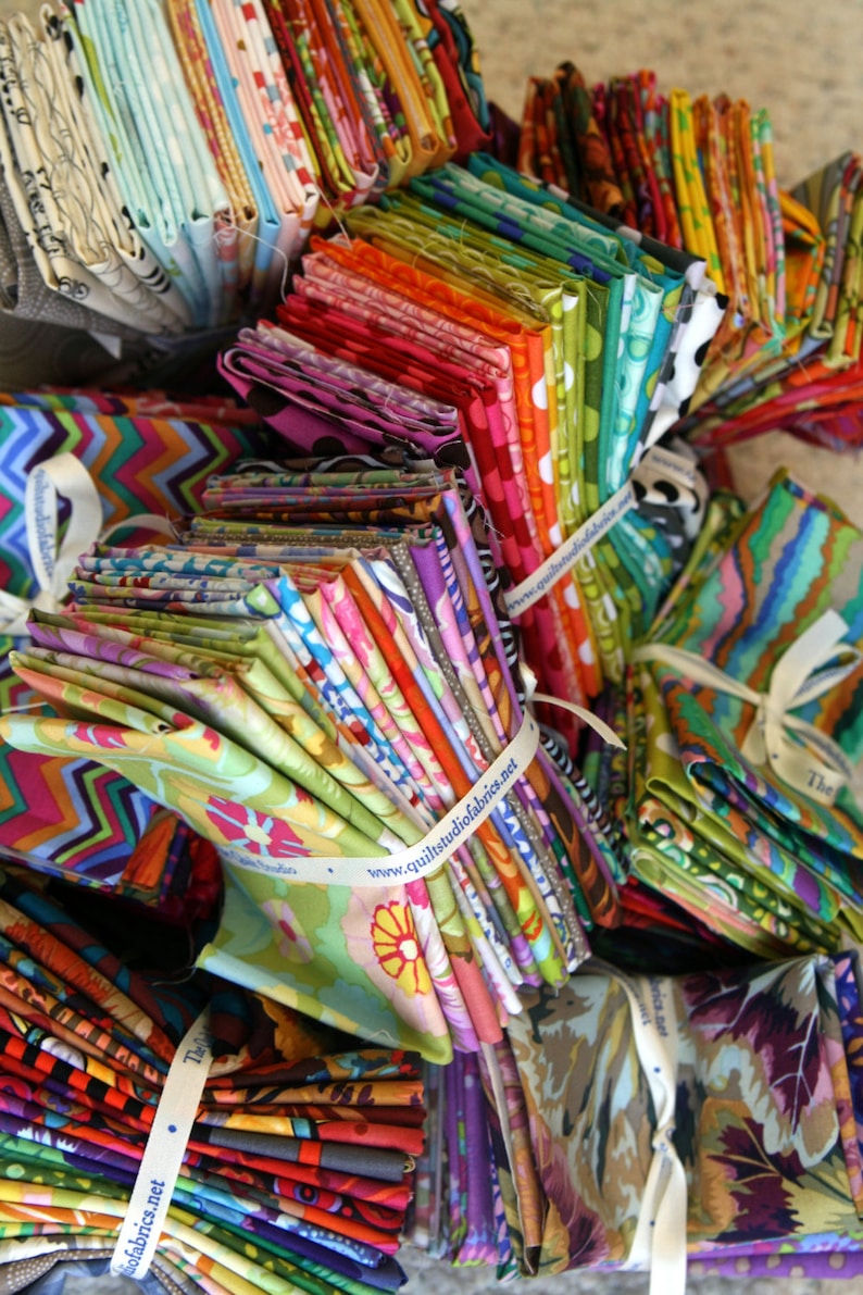May include: A large stack of colorful fabric scraps with various patterns and designs. The fabrics are arranged in a pile and tied with white ribbon. The ribbon has the text "www.quiltsudiofabrics.net" printed on it.