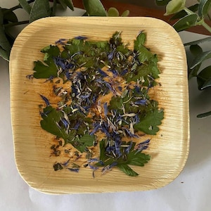 May include: A square, light brown wooden plate filled with green herbs and blue flower petals. The plate is surrounded by green leaves and sits on a white surface.