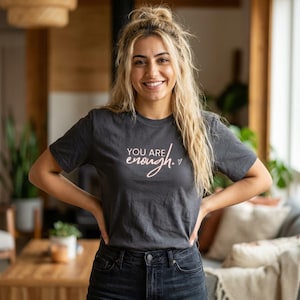 May include: Dark grey crew neck t-shirt featuring the text "YOU ARE enough." in light pink cursive. The shirt is being worn by a person. The background shows a living room with plants and a sofa, creating a relaxed atmosphere.