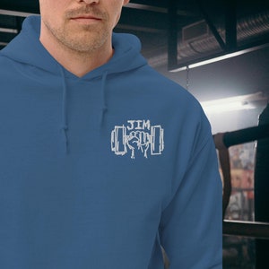 Könnte beinhalten: Ein blaues Kapuzenpullover mit einem weißen gestickten Design einer Faust, die eine Hantel hält, mit dem Wort "JIM" darüber.