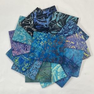 May include: A collection of folded fabric squares in various shades of blue, teal, and purple. Each square features a unique batik-style pattern of leaves, flowers, and abstract designs. The arrangement creates a circular, fan-like display.