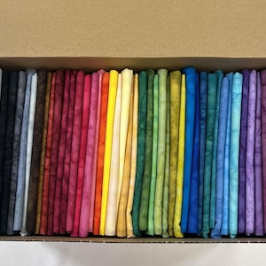 May include: A cardboard box filled with a collection of folded fabric strips arranged in a spectrum of colors. The fabric pieces feature a mottled, batik-style pattern and are organized sequentially from black and gray to red, yellow, green, blue, and purple.