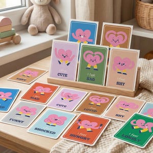 May include: A collection of emotion cards with a pink heart character. Each card has a different emotion label, such as "Cute," "Sad," and "Lonely." The cards are displayed on a wooden surface.