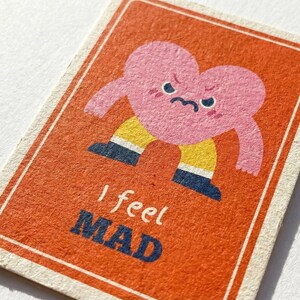 May include: A rectangular card with an orange background and a pink heart character with a grumpy face. The heart has arms and is wearing yellow pants and blue shoes. The text "I feel MAD" is printed in blue and white.