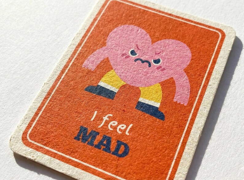 May include: A rectangular card with an orange background and a pink heart character with a grumpy face. The heart has arms and is wearing yellow pants and blue shoes. The text "I feel MAD" is printed in blue and white.