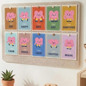May include: A felt board displays ten colorful cards, each with a cartoon heart character expressing different emotions. The cards are labeled with feelings like "Shy," "Happy," and "Brave." A small potted succulent and a woven basket are visible.