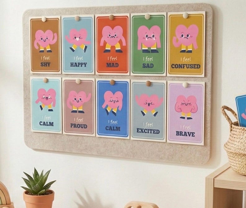 May include: A felt board displays ten colorful cards, each with a cartoon heart character expressing different emotions. The cards are labeled with feelings like "Shy," "Happy," and "Brave." A small potted succulent and a woven basket are visible.