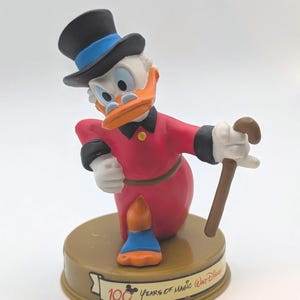 May include: A figurine of Scrooge McDuck, a cartoon duck, wearing a red coat, black top hat with a blue band, and holding a cane. The figurine is on a gold-colored base with the text "100 Years of Magic Walt Disney".